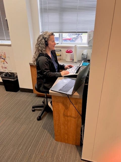 A team member working at a hearing testing workstation beside the sound booth at Sense of Hearing Welland