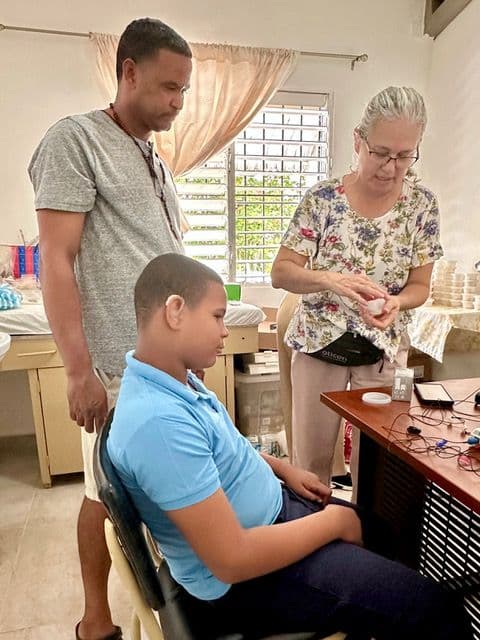 Two Perry-Medico clinicians prepare a hearing aid for fitting while a young patient waits in the chair