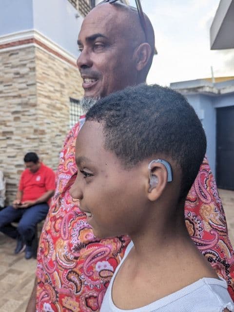 A young boy in Yamasa wearing a behind-the-ear hearing aid, standing with a smiling adult behind him outside the clinic