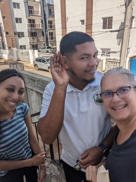 A young man in a school polo smiles and cups his hand to his newly fit hearing aid, posing with two Perry-Medico team members